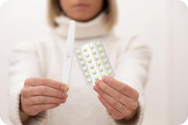 A woman in a cream jumper holding a pregnancy test and a blister pack of contraceptive pills
