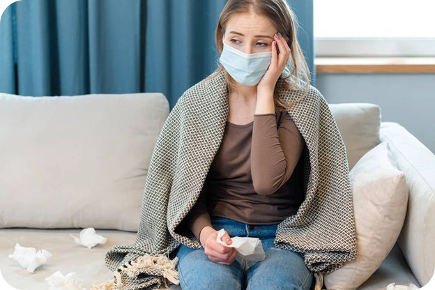 A woman wrapped in a blanket on a sofa, wearing a face mask and holding a tissue
